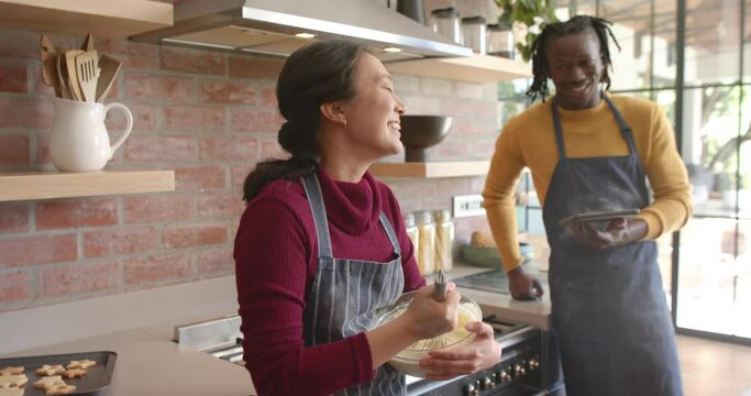 Happy diverse couple in aprons using tablet, mixing dough and talking in kitchen, slow motion