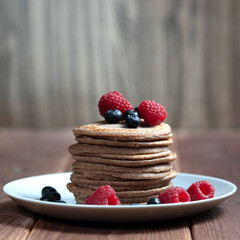 A stack of pancakes with fresh raspberries and blueberries on a light plate on a wooden background