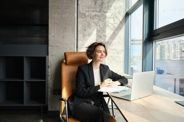 Portrait of young company employee, manager sitting in suit in her office, working on laptop,...