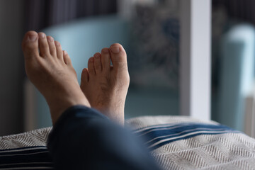 Closeup of a man's feet, relaxing on a bed in a hotel room. In the background is an upholstered...