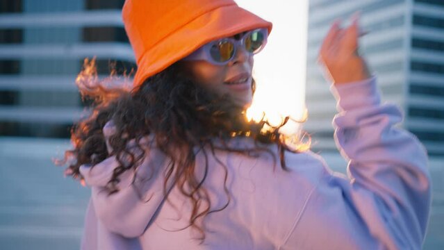 Fashion woman in trendy sunglasses and orange bucket hat dancing outside looking at camera. Outdoor portrait female dancer performing dance with freestyle moves. Funny stylish multiracial hipster girl