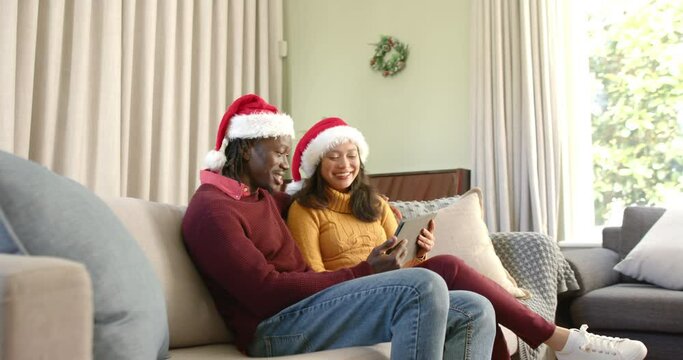 Happy diverse couple in christmas hats using tablet on couch at home, copy space, slow motion