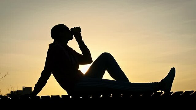 Young woman drinks warm coffee at a beautiful sunset sky in slow motion. A female silhouette in setting sun rays sits on a parapet in a park with coffee in her hands and enjoys life. Healthy lifestyle