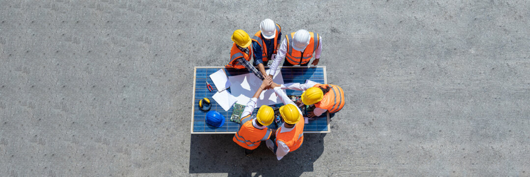 Top view of engineers team, male and female put hands on top over blueprint, solar photovoltaic equipment, road and wind turbine business important infrastructure on solar panel at construction site.
