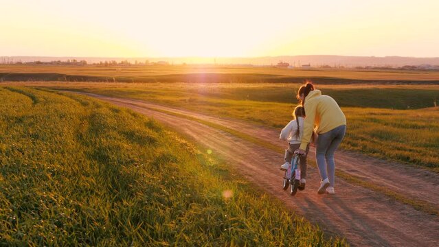 Loving mother teaches little girl to ride bicycle playing in sunset field