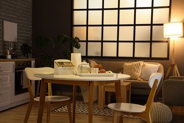 Interior of modern kitchen with glowing lamps, dining table and grey sofa at evening