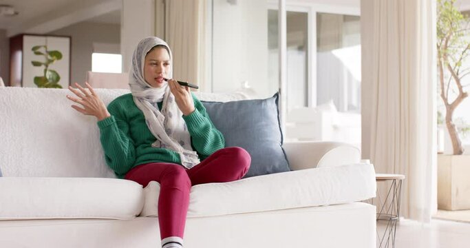 Biracial woman in hijab using smartphone on sofa at home with copy space, slow motion