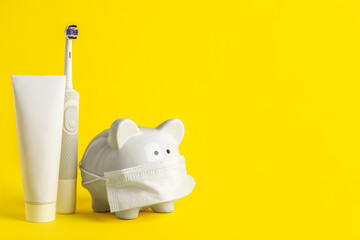 Piggy bank with medical mask, toothpaste and electric toothbrush on yellow background