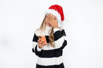 Beautiful hispanic woman wearing christmas hat and striped knitted sweater looking at smart phone feeling sad holding hand on face.