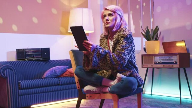 Horizontal shot. Hipster woman sitting and holding tablet. Stylish female chilling at disco styled room. Woman holding gadget and surfing on internet. Pink hair woman relaxing in retro vintage room.