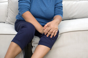 A distressed senior woman experiencing sharp knee pain seated on a comfy sofa, her hand tenderly resting on the sore spot. - obrazy, fototapety, plakaty