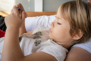 Cute little boy using smartphone, lying on cozy couch, relaxing on weekend, enjoying leisure time, interested kid looking at phone screen, holding mobile device, playing game, watching cartoons