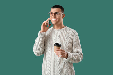 Young man in knitted sweater with cup of coffee talking by mobile phone on green background