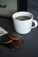 White cup with coffee next to cookies on a wooden tray and a music speaker on a gray table