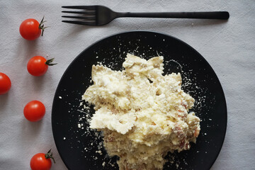 Pasta carbonara with parmesan cheese on a black plate with a black fork next to cherry tomatoes on a gray tablecloth
