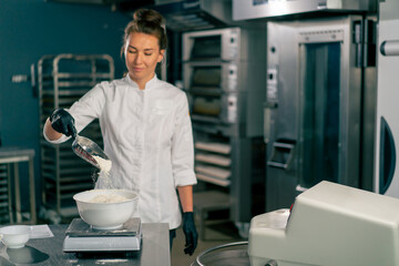 female baker pours wheat flour into mixer for kneading dough for preparing fresh baked goods