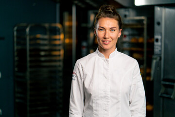 Portrait of a smiling female chef baker in a professional uniform in a bakery against background of an oven and fresh baked goods