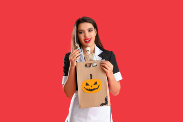 Young woman dressed for Halloween as maid with gift bag and skeleton on red background