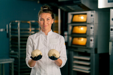 smiling female baker stands in professional kitchen against the background of ovens and holds...