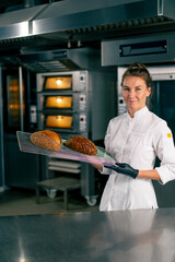 smiling female chef standing with freshly baked loaves of bread in the kitchen of a bakery and looking at camera