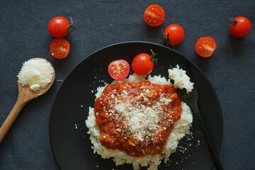 Rice with tomato sauce, meatballs and parmesan on a black plate next to a fork and cherry tomatoes on a gray tablecloth