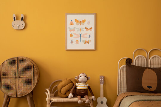 Aesthetic composition of child room interior with mock up poster frame, yellow wall, plush toys, monkey, rattan sideboard, guitar, brown bedding and personal accessories. Home decor. Template.