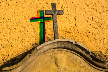 Cross on a tombstone at a yellow wall with shadow in rainbow colors