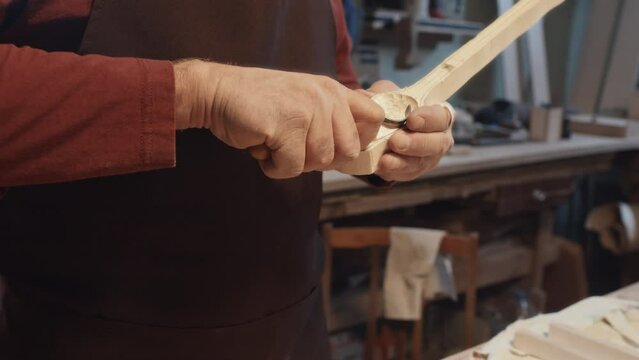Selective focus medium close-up of hands of unrecognizable Caucasian elderly man wearing apron using chisel carving out new cutlery