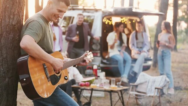 A group of friends on vacation in the forest on a warm summer evening. A picnic with a car and a fire. the guy plays the guitar