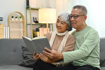 Fototapeta na wymiar Beautiful senior couple embracing and reading book on sofa while spending time together at home