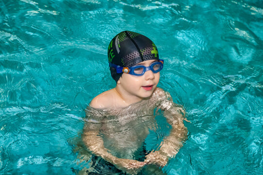 Portrait of child boy swimmer in swimming goggles and swim hat in water pool workout, pensive looking. Kid 5 year old in exercising in pool. Sports children training concept. Copy ad text space