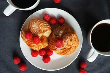 Cakes with cream and raspberries on a white plate next to a cup of coffee and a knife on a dark background