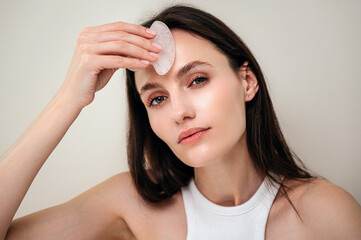 Young woman massages forehead with a quartz gua sha scraper - obrazy, fototapety, plakaty