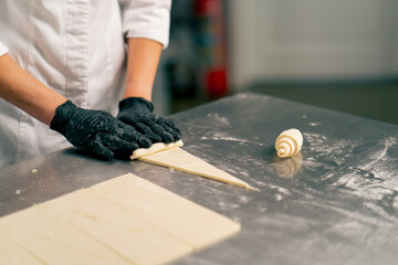 Close-up shot of chef's hand in gloves rolling raw cut dough into croissant shapes for baking