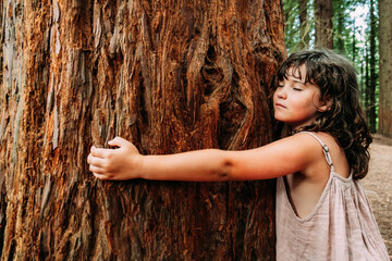 Calm little girl embracing huge old tree trunk in forest