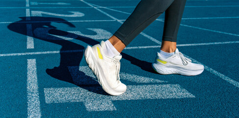 Detail of legs and sports shoes of a young female runner, prepared in a position with her legs and feet flexed to do sprint or a race on a blue athletics track. Runner's shadow projected on the track.