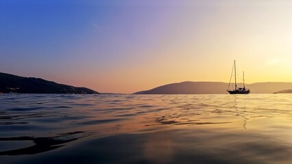 View from the calm sea surface on yacht silhouette against sunset sky and mountains.