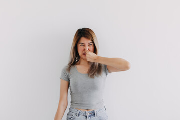 Unhappy Asian Thai woman wear grey, squeezing, pinching and holding nose, blocking bad smell, looking at camera smelly, isolated on white background wall.