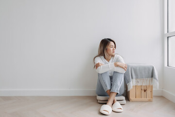 Asian Thai woman sitting on floor and hugging knees, looking at the window, feeling sad and worried, thinking something lonely, absent-minded at apartment in winter. 
