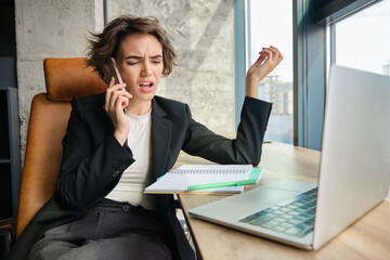 Portrait of angry businesswoman, sitting in office with laptop, having an argument over the phone