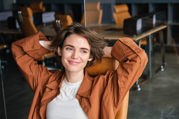 Close up portrait of woman at workplace, digital nomad sits back and relaxed in an office, resting after productive work day, smiling pleased