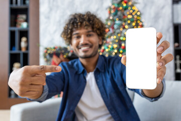 Man sitting on a sofa in living room on winter day Christmas. Hispanic shows an empty white phone screen to the camera, presents an application on a smartphone, celebrates the New Year near tree.