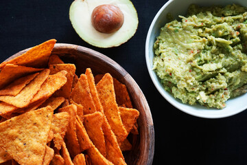 Plates with nachos and guacamole next to avocado on black background