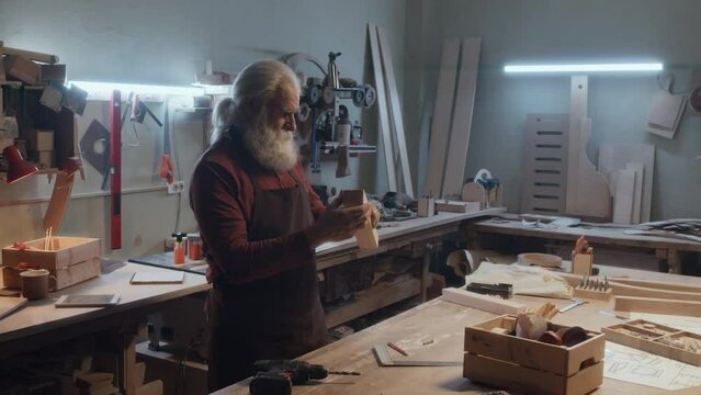 Zoom in of senior Caicasian carpenter making hole in piece of wood with drill and checking his work
