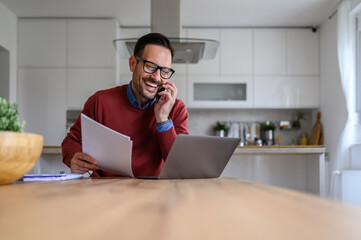 Happy young businessman holding documents and talking over smart phone while working from home