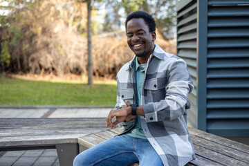Cheerful young black man sitting on bench with watch in daytime