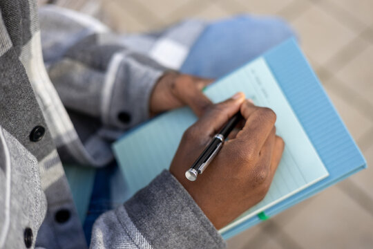 Unrecognizable black man sitting with pen and notebook in daytime