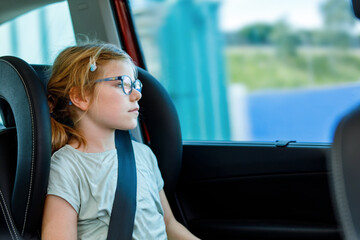 Little Preschool Girl Sitting in Her Car Seat. Happy Child with Eyeglasses looking out of the window. Bored kid on the Way to Family Vacations during Traffic Jam.