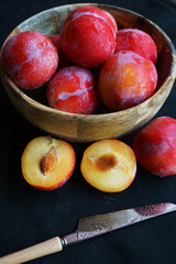 Ripe plums in a bowl next to sliced ​​plums and a knife on a dark tablecloth