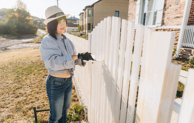 beautiful asian woman paints a wooden fence with white paint.